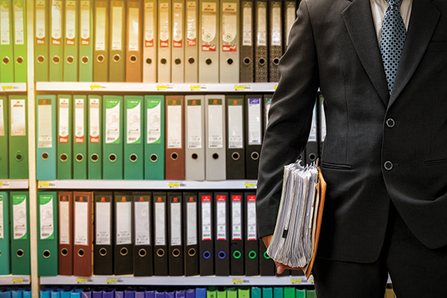 Person in a suit pictured from the neck down standing in front of a shelf of legal files, holding a stack of legal papers.