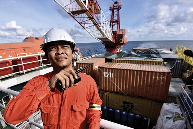 A man in a jumpsuit and hardhat on a shipping boat, holding a walkie talkie. Behind him is a crane and shipping containers.