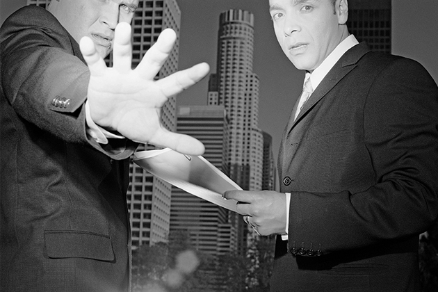 Black and white photo of two men in suits. One holds documents and the other puts out his hand to block the camera, implying that their conversation is business-related and illicit.