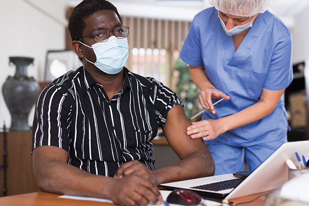A man wearing a COVID mask, sitting in front of an open laptop receives a shot in his arm from a nurse wearing scrubs and a COVID mask.