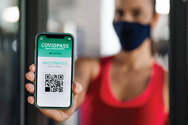 A woman wearing a COVID mask holds up a cell phone showing an app called COVIDPASS with the text 