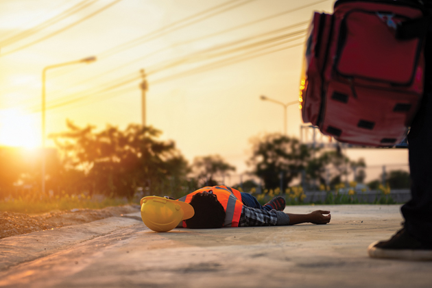 A worker lying prone on the ground with a hardhat sitting next to them. In the foreground, a person with a first aid pack approaches.