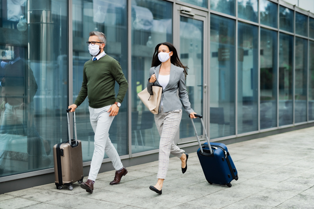 A man and woman walk with rolling suitcases while wearing surgical masks to protect against COVID-19.