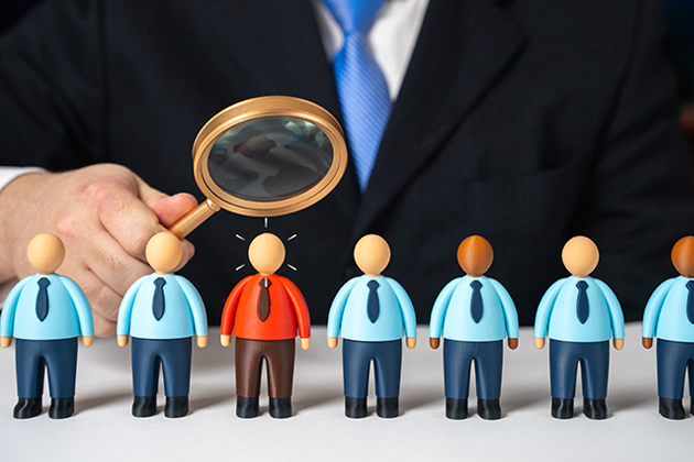 Businessperson use magnifying glass to examine red-shirted employee standing out in in row of employees wearing blue shirts