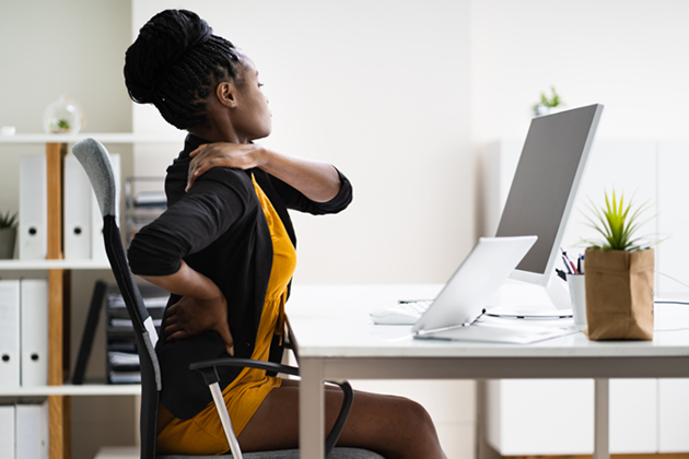 Woman sitting at desk in home office facing multiple computers and massaging her shoulder and back in pain.