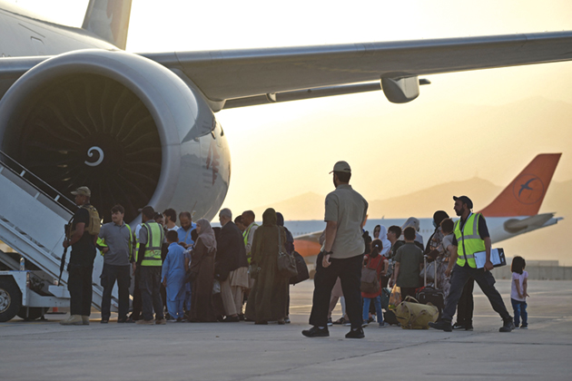 A group of families waiting to board an airplane, guided by men in bright yellow vests and guarded by a man with a gun.