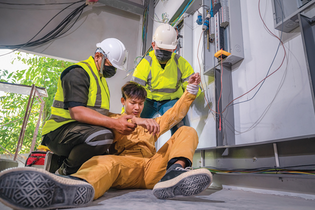 Two men in hardhats, bright yellow vests and COVID masks hold a third man, who is collapsed on the ground and whose hand is near some exposed wires.