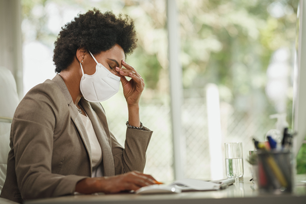 A Black woman wearing a business suit sits at a desk in front of a computer. She is wearing a COVID mask, has her eyes closed, and is holding the bridge of her nose with one hand, showing exhaustion or frustration.