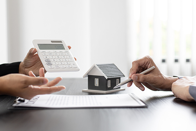 Two people's hands across a desk: One holds a calculator and the other a pen. In front of the person with the pen is a clipboard and a small model of a house.