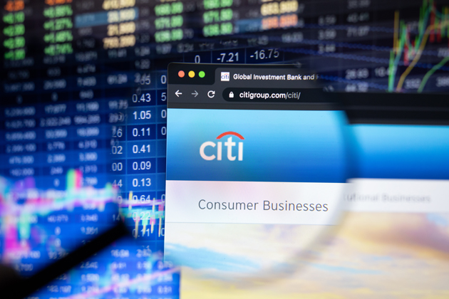 A magnifying glass in front of a computer screen showing a Citi Consumer Business logo. In the background is a screen showing stock market numbers.