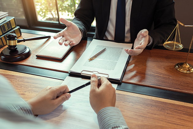 Two people across from each other across a desk, pictured from the torso down. One is holding a pen, the other has a clipboard in front of him with the scales of justice and a gavel, implying that the first person is facing legal consequences.