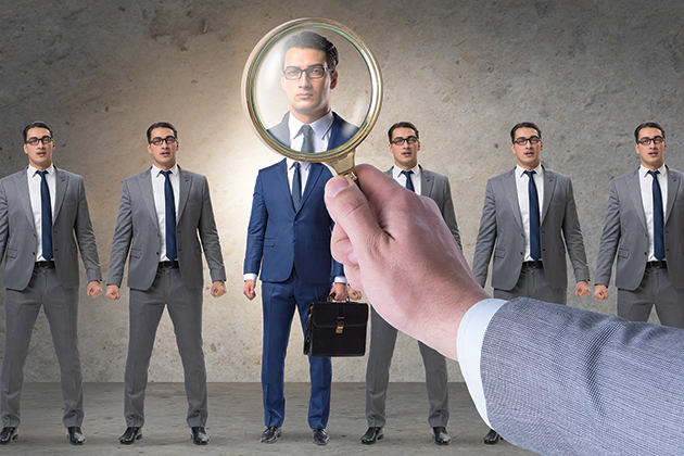 A line of identical men in grey suits wearing glasses. In the foreground, a hand holds a magnifying glass over the man in the center of the line, who is the only one wearing a blue suit and carrying a briefcase. His face is shown in the magnifying glass.