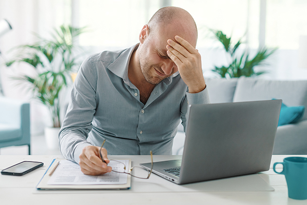 A man in his home sitting in front of an open laptop and notepad. He has taken off his glasses and is holding his forehead and grimacing.