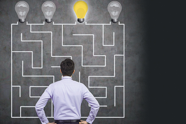 A man in business attire faces a wall, on which is a maze drawn in chalk leading to four lightbulbs, one of which is lit up.