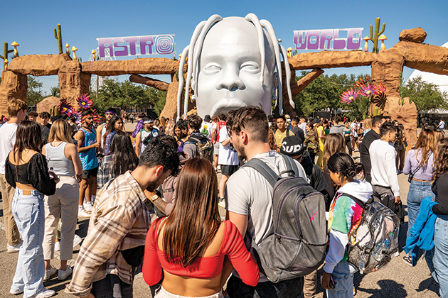 Crowds of concert attendees at the Astro World music festival. In the background, there is a giant sculpture of Travis Scott's head and a sign for the festival.