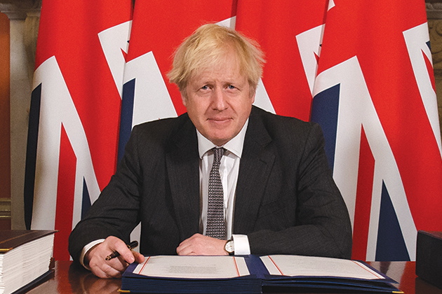 UK Prime Minister Boris Johnson sits at a table in front of four UK flags. He holds a pen and in front of him is an open folder.