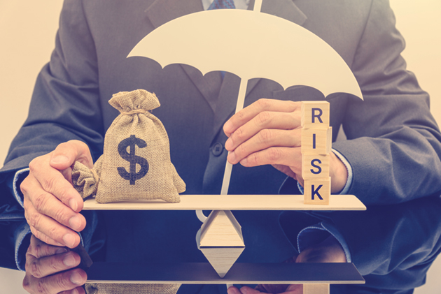 A person in a suit holding an umbrella over a scale that is balancing a bag of money and blocks spelling the word 