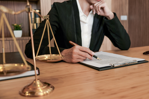 A person sits at a desk putting a pen to a form on a clipboard, with the scales of justice on the desk in the foreground.