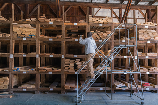 A man inspects a stack of cut wood while standing on a ladder.