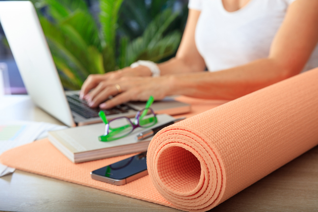 A woman typing on a laptop at a desk with a peach-colored yoga mat on the desk in the foreground.