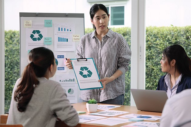 Three people in meeting with one holding up clipboard with recycle sign