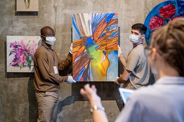 Two mean wearing masks and gloves hanging an abstract paining on a concrete wall while a third person observes.